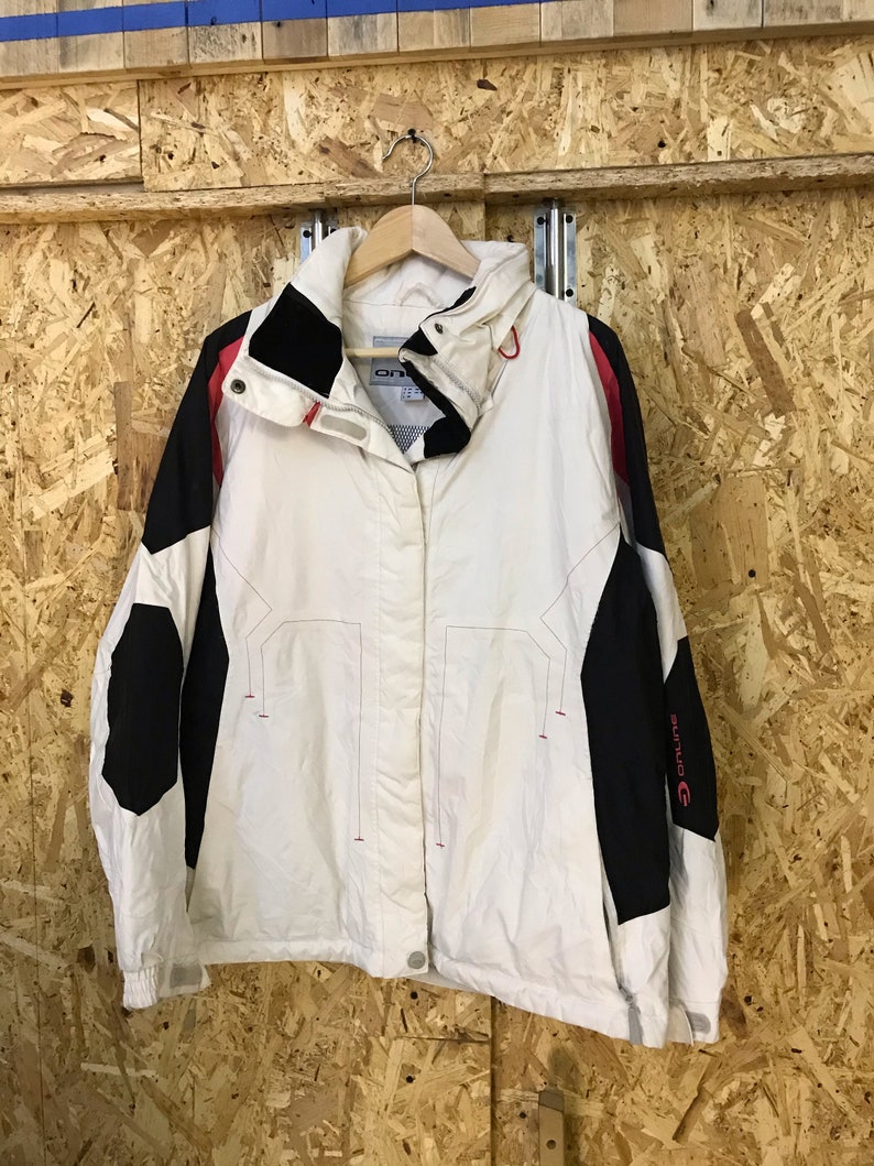 Puede incluir: Chaqueta blanca y negra con detalles rojos, colgada de una percha de madera. La chaqueta tiene un cuello alto y cierre de botones. Las mangas son negras con un logotipo rojo. La chaqueta es de material ligero.