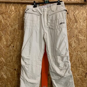 Puede incluir: Pantalones de nieve color crema con m&uacute;ltiples bolsillos y detalles cosidos. Los pantalones tienen cremallera, cierre de bot&oacute;n y un peque&ntilde;o logotipo en la pierna derecha. Los pantalones est&aacute;n colgados de una percha negra.