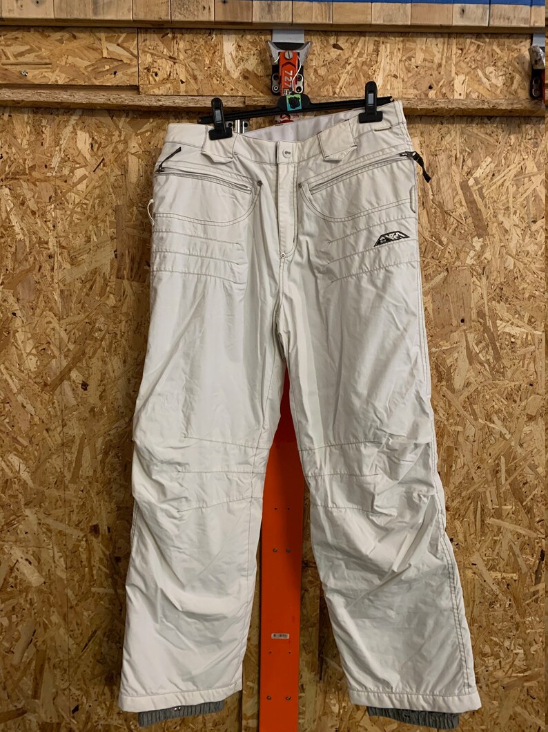 Puede incluir: Pantalones de nieve color crema con m&uacute;ltiples bolsillos y detalles cosidos. Los pantalones tienen cremallera, cierre de bot&oacute;n y un peque&ntilde;o logotipo en la pierna derecha. Los pantalones est&aacute;n colgados de una percha negra.