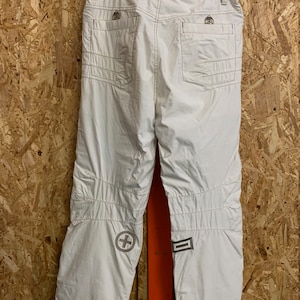 Puede incluir: Pantalones de nieve blancos con m&uacute;ltiples bolsillos y rodillas reforzadas. Los pantalones tienen una cruz y un dise&ntilde;o rectangular en la parte inferior de las piernas. Las costuras son de color marr&oacute;n claro. Los pantalones cuelgan de una percha negra.