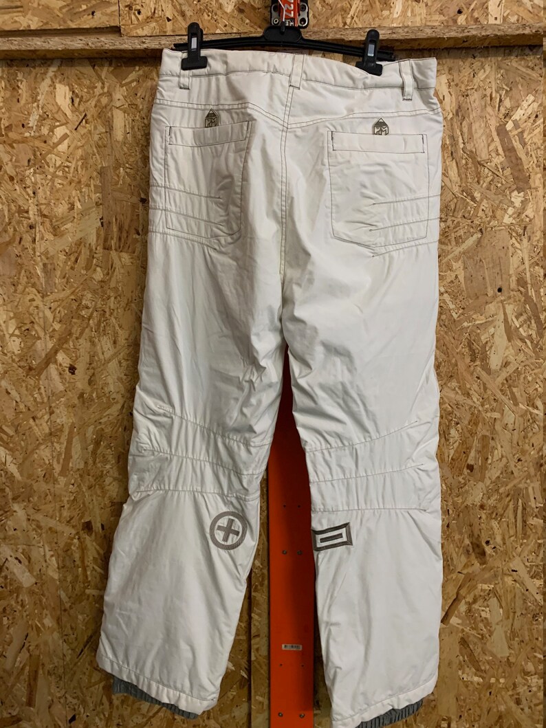 Puede incluir: Pantalones de nieve blancos con m&uacute;ltiples bolsillos y rodillas reforzadas. Los pantalones tienen una cruz y un dise&ntilde;o rectangular en la parte inferior de las piernas. Las costuras son de color marr&oacute;n claro. Los pantalones cuelgan de una percha negra.