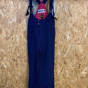 May include: Navy blue ski bib overalls with adjustable straps and a red interior lining. The bib features the brand name "BAILO" and a "GORE-TEX" label. The overalls are hanging on a black hanger against a wood-paneled wall.
