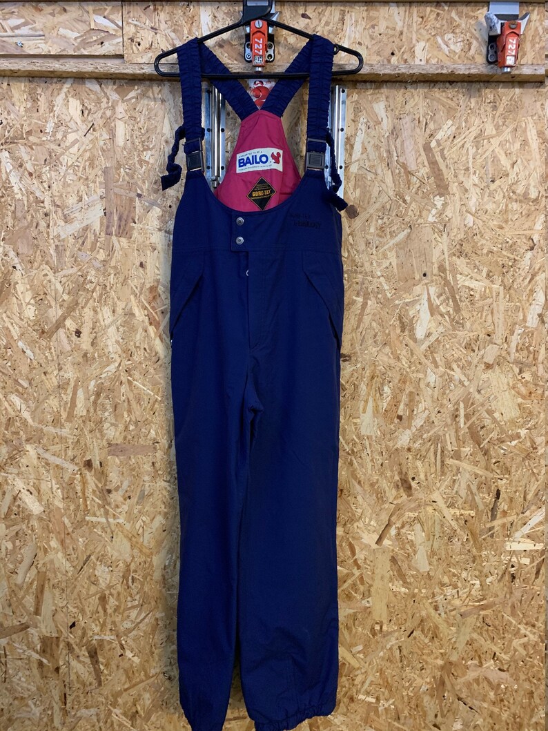 May include: Navy blue ski bib overalls with adjustable straps and a red interior lining. The bib features the brand name "BAILO" and a "GORE-TEX" label. The overalls are hanging on a black hanger against a wood-paneled wall.