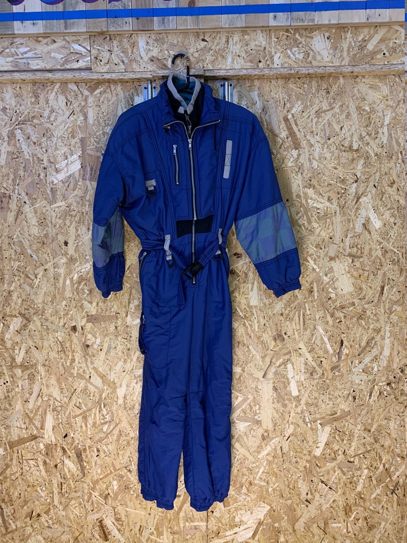 May include: A blue vintage ski suit with a black belt and gray accents on the sleeves. The one-piece suit has a zipper closure and is hanging on a metal hanger. The suit is made of a water-resistant material.