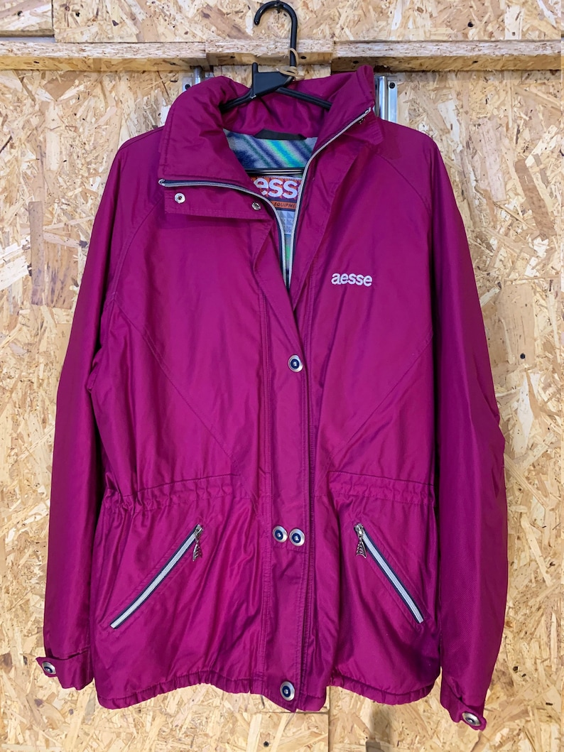 Pode incluir: Casaco magenta com gola alta e cintura ajust&aacute;vel por cord&atilde;o. O casaco tem fecho de correr e dois bolsos frontais com z&iacute;peres. O nome da marca "aesse" &eacute; bordado no peito. Pendurado num cabide preto sobre um fundo de madeira.