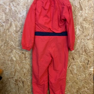 May include: A bright red one-piece ski suit with a black waist band. The suit has a high collar and long sleeves. The suit is hanging on a black hanger against a wooden background.