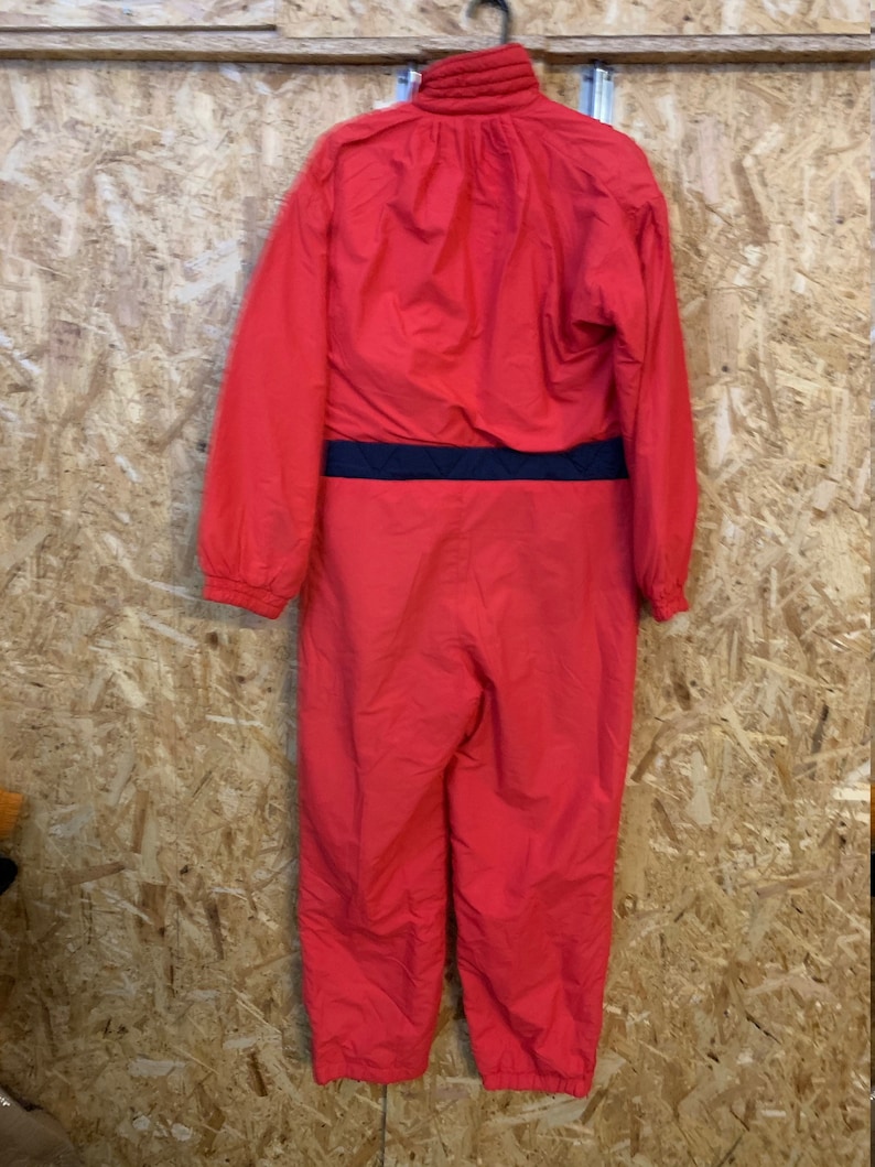 May include: A bright red one-piece ski suit with a black waist band. The suit has a high collar and long sleeves. The suit is hanging on a black hanger against a wooden background.