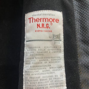 May include: Close-up of a white tag with the text "Thermore N.R.G. ENERGY SAVING" in red. The tag also includes text in multiple languages describing thermal insulation, warmth, comfort, and freedom of movement. The tag is attached to a black garment.