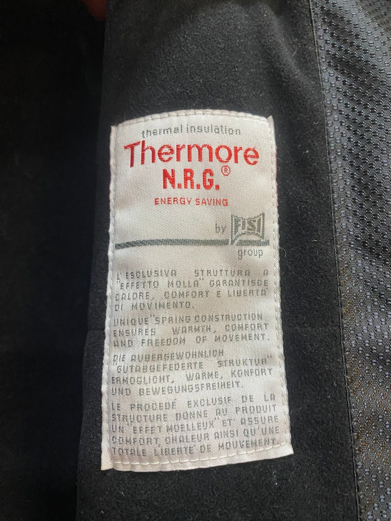 May include: Close-up of a white tag with the text "Thermore N.R.G. ENERGY SAVING" in red. The tag also includes text in multiple languages describing thermal insulation, warmth, comfort, and freedom of movement. The tag is attached to a black garment.