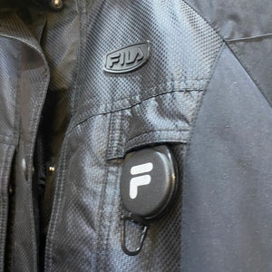 May include: Black FILA jacket with a textured pattern and a small logo on the chest. The jacket has a pocket with a retractable reel featuring the FILA logo. The jacket is made of a durable, weather-resistant material.