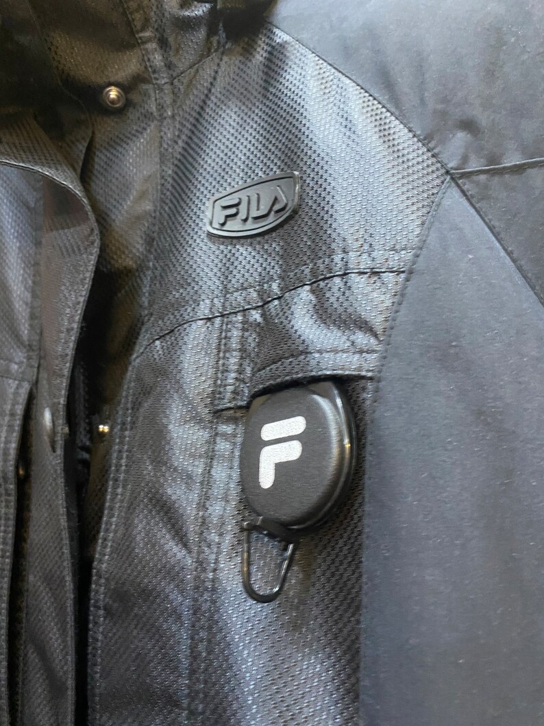 May include: Black FILA jacket with a textured pattern and a small logo on the chest. The jacket has a pocket with a retractable reel featuring the FILA logo. The jacket is made of a durable, weather-resistant material.