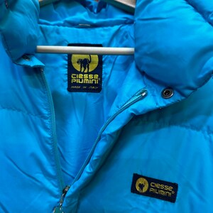 May include: A close-up of a bright turquoise puffer jacket with a high collar and a zipper closure. The jacket features a label with a black cat logo and the text "Ciesse Piumini, Made in Italy". The jacket is hanging on a wooden hanger.