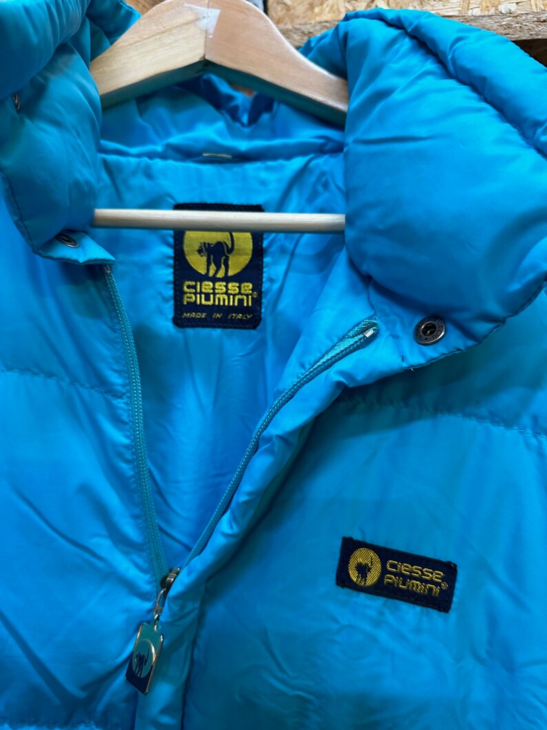 May include: A close-up of a bright turquoise puffer jacket with a high collar and a zipper closure. The jacket features a label with a black cat logo and the text "Ciesse Piumini, Made in Italy". The jacket is hanging on a wooden hanger.