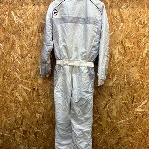 May include: A white and gray vintage ski suit with a collared neckline and a belt at the waist. The suit has a light gray stripe across the chest and a logo on the upper left chest. The suit is hanging against a wooden background.