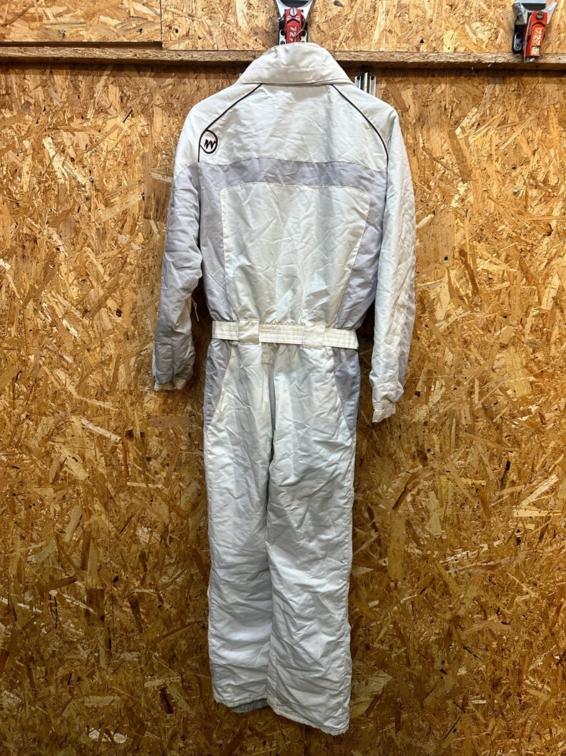 May include: A white and gray vintage ski suit with a collared neckline and a belt at the waist. The suit has a light gray stripe across the chest and a logo on the upper left chest. The suit is hanging against a wooden background.