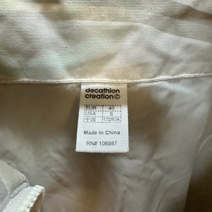 May include: Close-up of a tag from a cream-colored jacket. The tag reads "decathlon creation" and includes size information: EUR 40, USA S, and Chinese size 170/92A. The tag also states "Made in China" and includes the RN# 106987.