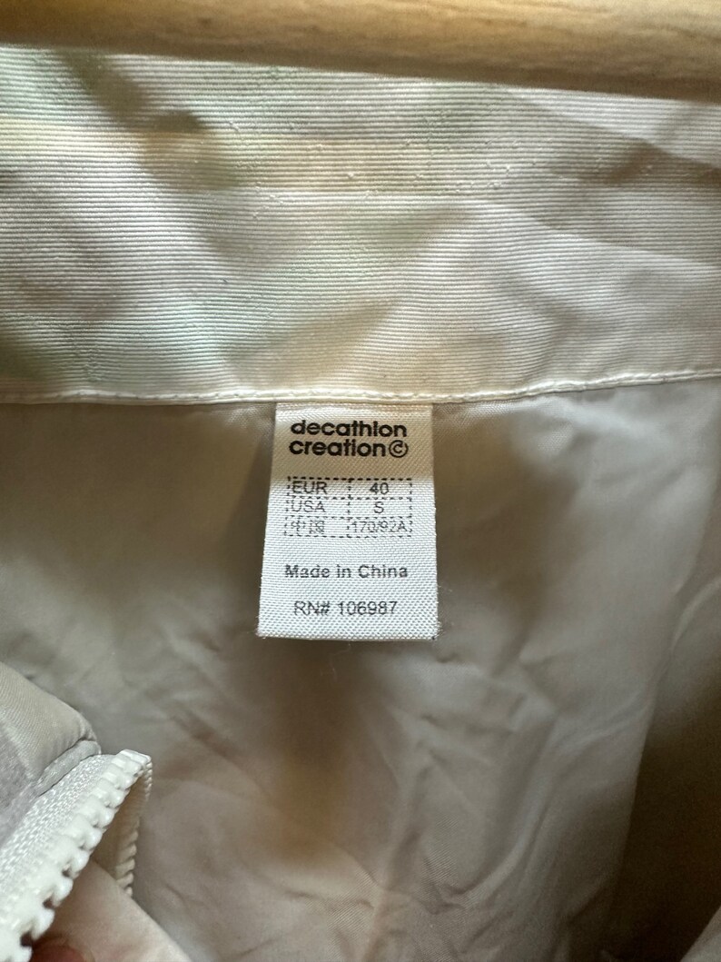 May include: Close-up of a tag from a cream-colored jacket. The tag reads "decathlon creation" and includes size information: EUR 40, USA S, and Chinese size 170/92A. The tag also states "Made in China" and includes the RN# 106987.