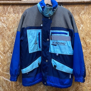 Könnte beinhalten: Eine blau-graue Racing-Jacke mit mehreren Taschen und dem Wort "RACING" auf der Vorderseite. Die Jacke hat einen hohen Kragen und einen Reißverschluss. Die Jacke besteht aus strapazierfähigem Material und ist für Outdoor-Aktivitäten konzipiert.