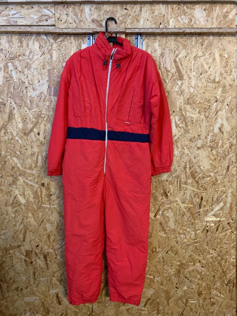 May include: A red one-piece snowsuit with a navy blue stripe across the midsection. The snowsuit has a white zipper, a high collar, and is hanging on a black hanger. The garment is made of a lightweight material.