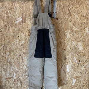 May include: Tan and black snow bib overalls hanging on a black hanger. The bibs have adjustable straps and a black panel on the front. The legs have zippers on the sides. The background is a light brown wood.