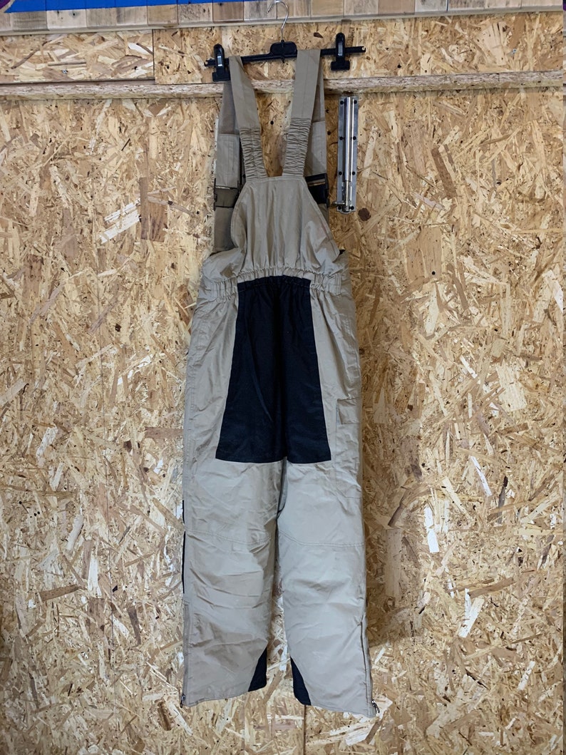 May include: Tan and black snow bib overalls hanging on a black hanger. The bibs have adjustable straps and a black panel on the front. The legs have zippers on the sides. The background is a light brown wood.