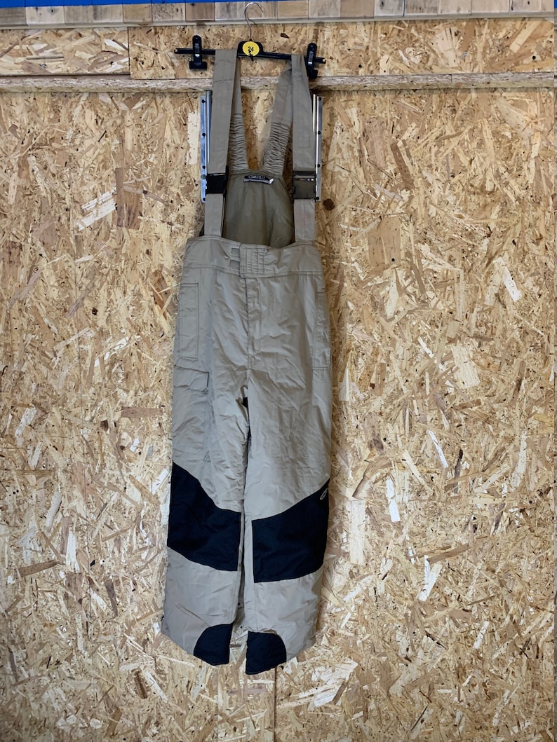 May include: Tan snow bib overalls with black reinforced knees hang from a black hanger. The bibs have adjustable straps and pockets. The background is a light brown wood panel.