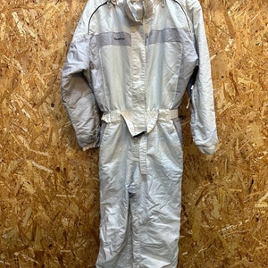 May include: A light gray one-piece ski suit with a hood, belt, and the brand name "Quechua" on the chest. The suit has a zipper closure and black trim. The suit is designed for cold weather activities.