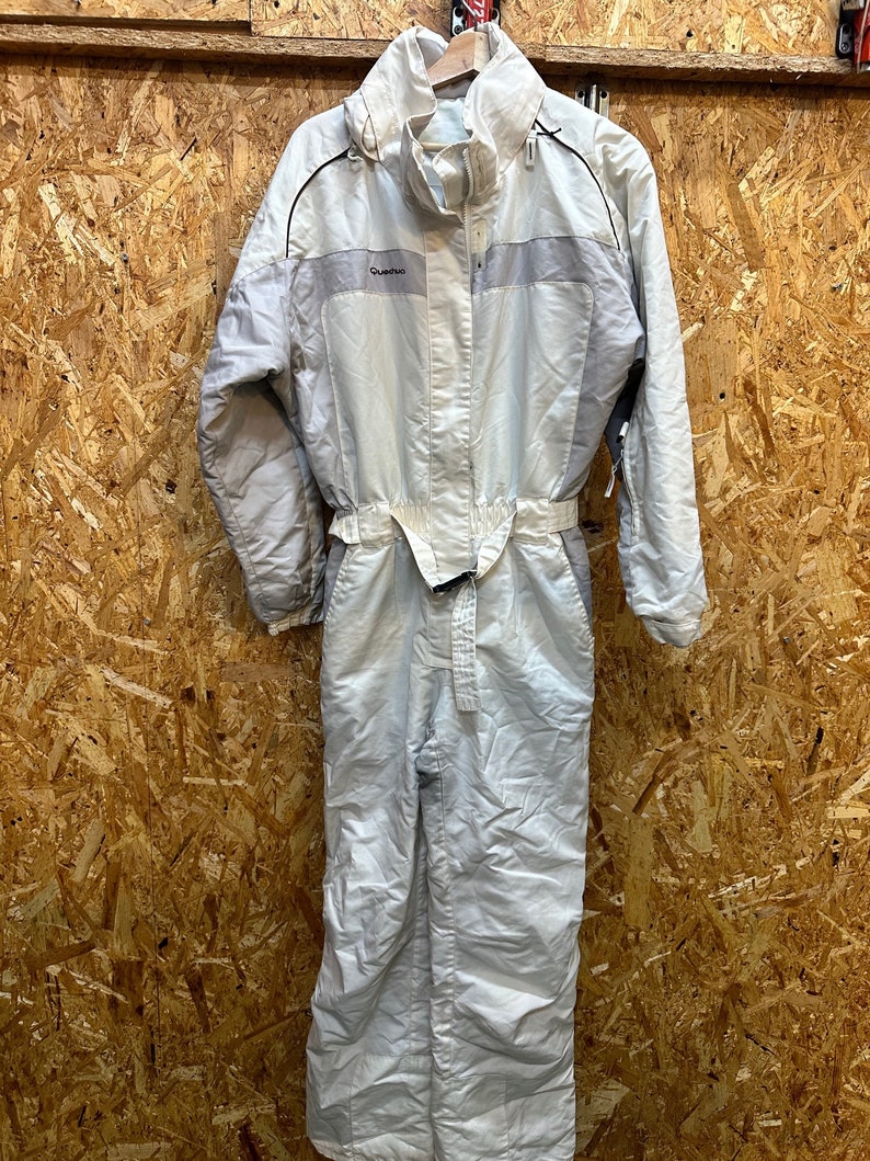 May include: A light gray one-piece ski suit with a hood, belt, and the brand name "Quechua" on the chest. The suit has a zipper closure and black trim. The suit is designed for cold weather activities.