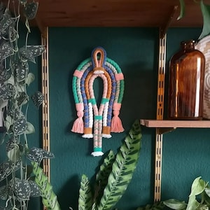 May include: A colorful macrame wall hanging with a rainbow design and tassels. The hanging is made with different colored cords and is displayed on a green wall.