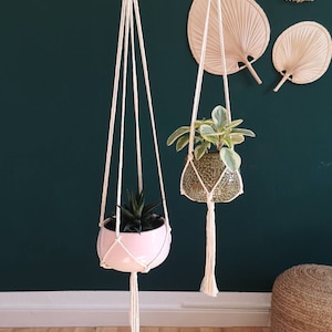 Puede incluir: Dos maceteros colgantes de macram&eacute; con anillos de madera, cada uno con una planta en maceta. Los maceteros est&aacute;n hechos de cuerda de algod&oacute;n natural y tienen borlas largas. Las plantas est&aacute;n en macetas de cer&aacute;mica rosa y verde.