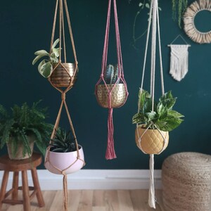 Puede incluir: Tres maceteros colgantes de macram&eacute; con cordones y borlas de diferentes colores. Cada macetero sostiene una planta en maceta diferente en una maceta met&aacute;lica o de cer&aacute;mica.