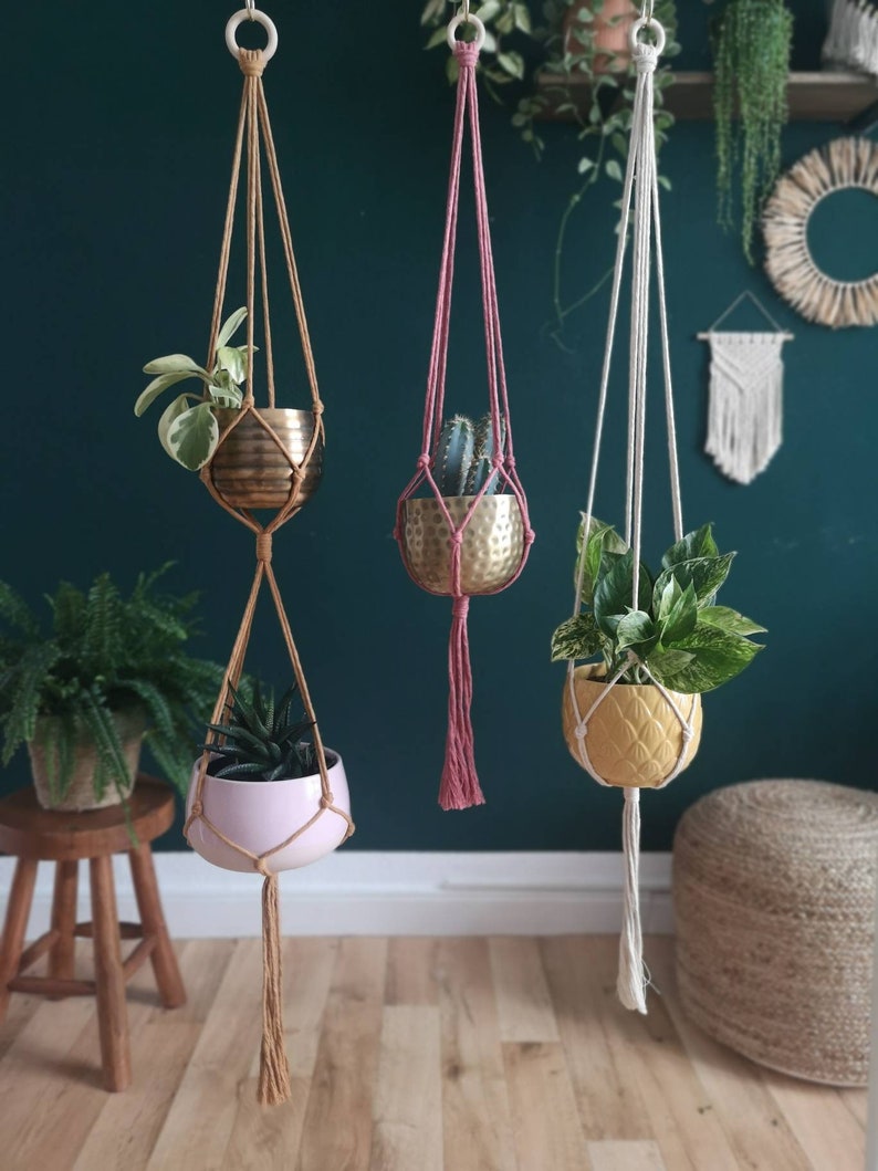 Puede incluir: Tres maceteros colgantes de macram&eacute; con cordones y borlas de diferentes colores. Cada macetero sostiene una planta en maceta diferente en una maceta met&aacute;lica o de cer&aacute;mica.