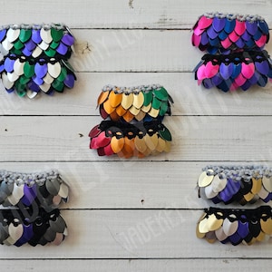 May include: Six pairs of colorful, layered, scale mail bracelets. Each bracelet is made of overlapping scales in a variety of colors, including purple, green, blue, pink, gold, and black. The bracelets are displayed on a white wooden surface.