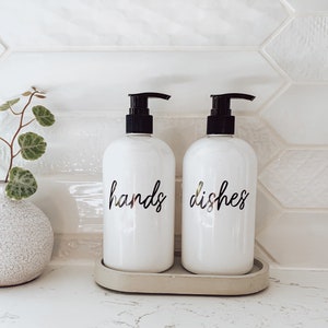 May include: Two white ceramic soap pumps with black tops and gold lettering. The pump on the left says "hands" and the pump on the right says "dishes". The pumps are sitting on a grey tray.
