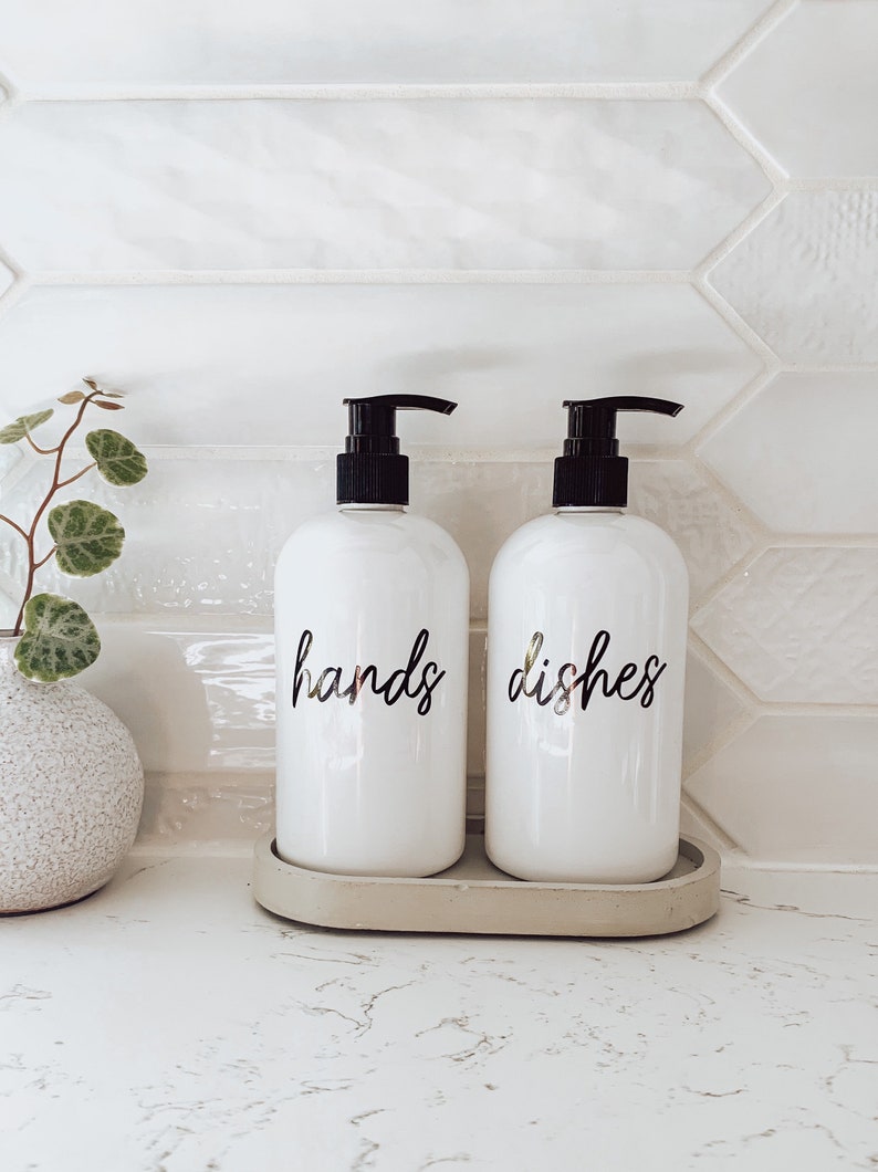 May include: Two white ceramic soap pumps with black tops and gold lettering. The pump on the left says "hands" and the pump on the right says "dishes". The pumps are sitting on a grey tray.