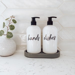 May include: Two white pump bottles with black pumps and labels that say "hands" and "dishes" on a grey oval tray. The bottles are on a white countertop with a white tiled backsplash.