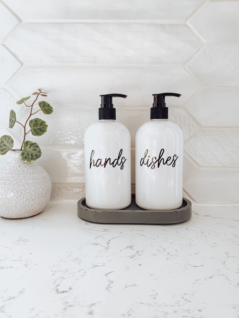 May include: Two white pump bottles with black pumps and labels that say "hands" and "dishes" on a grey oval tray. The bottles are on a white countertop with a white tiled backsplash.