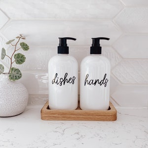 May include: Two white soap pumps with black tops and labels that say "dishes" and "hands" on a wooden tray.