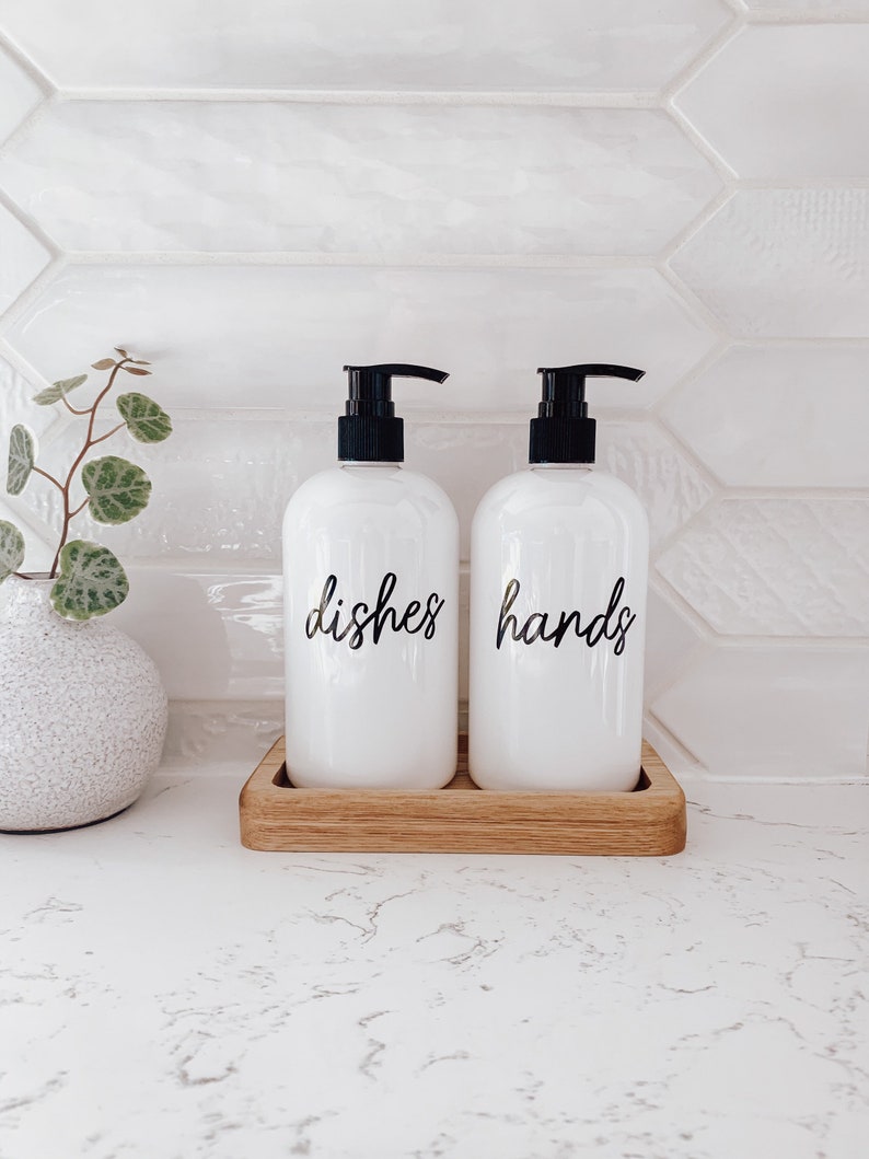 May include: Two white soap pumps with black tops and labels that say "dishes" and "hands" on a wooden tray.