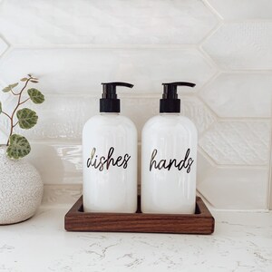 May include: Two white pump bottles with black pumps and labels that say "dishes" and "hands" on a wooden tray.