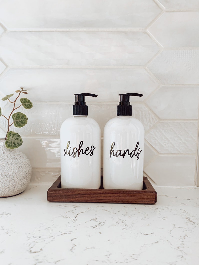 May include: Two white pump bottles with black pumps and labels that say "dishes" and "hands" on a wooden tray.