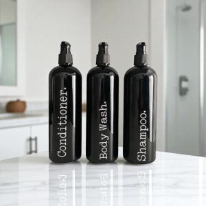 May include: Three black pump bottles with white labels reading "Conditioner," "Body Wash," and "Shampoo." They stand on a white, reflective surface. The bottles have black pumps, suitable for a bathroom setting.