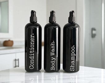 May include: Three black pump bottles labeled "Conditioner," "Body Wash," and "Shampoo" in white text. The bottles are arranged on a white, reflective surface, suggesting a bathroom setting. The bottles have black pumps.