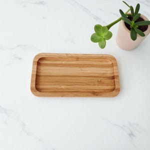 May include: A rectangular bamboo tray with rounded corners. The tray is light brown with visible wood grain. A small potted plant with green leaves sits in the upper right corner. The background is a white marble surface.