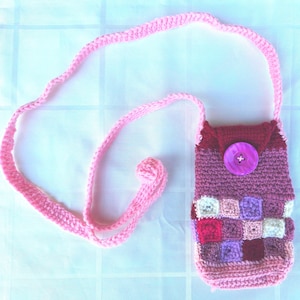 May include: A handmade crochet bag in shades of pink and purple, featuring a patchwork design on the front. The bag has a long, pink strap and a flap secured with a large, round button. The bag is designed to be worn over the shoulder.
