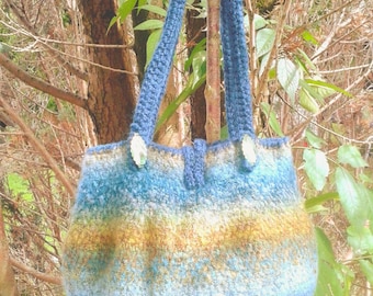 May include: A crocheted tote bag featuring a gradient of blue, teal, and golden-brown hues. The bag has two blue crocheted handles, two clear decorative accents at the base of the straps, and a row of small metallic beads along the bottom edge.