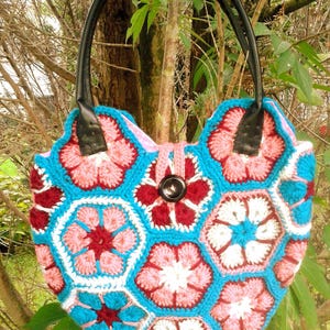 Può includere: Borsa a mano all'uncinetto fatta a mano con motivo floreale. La borsa presenta uno sfondo turchese con motivi floreali rosa, bianchi e bordeaux. Ha manici in pelle nera e una chiusura con bottone. Un accessorio unico e colorato.