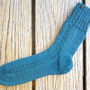 May include: A teal knitted sock featuring a vertical cable knit pattern running along the side, a ribbed cuff at the top, and a small dark brown button attached near the upper edge. The sock is constructed with a textured heel and toe section.