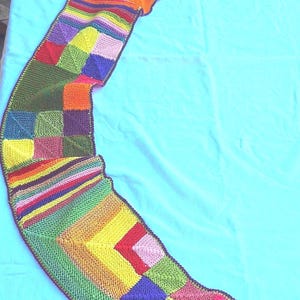 Puede incluir: Una colorida bufanda tejida a mano con un diseño de patchwork. La bufanda presenta cuadrados y rayas en varios colores, incluyendo rojo, amarillo, verde, morado y rosa. La bufanda mide aproximadamente 183 cm de largo.