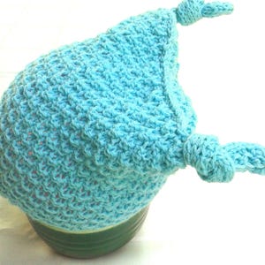 May include: A light blue crocheted hat with two knotted ties. The hat is sitting on a green surface.